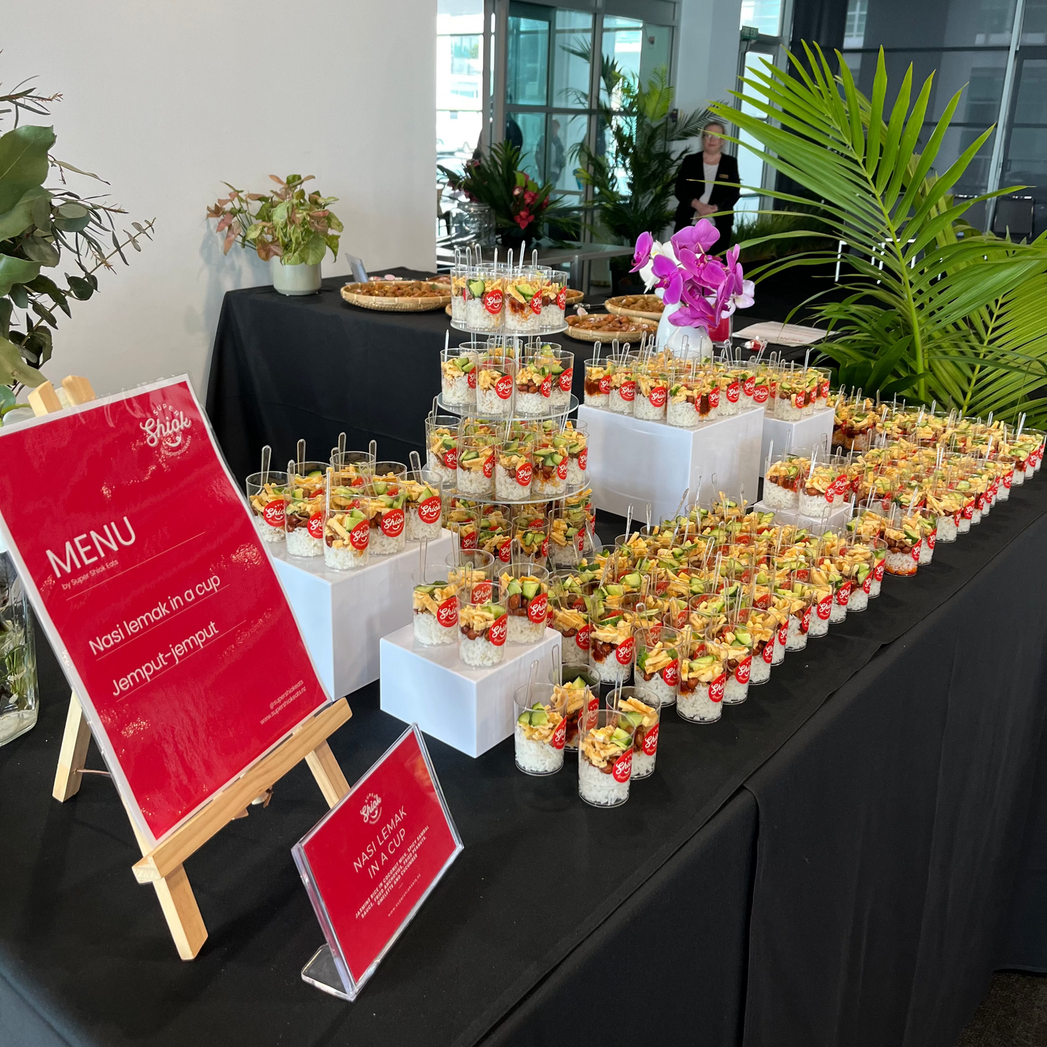 A catering display table covered with a black tablecloth, filled with stacks of mini appetizers and small dessert cups. Red signboards on easels display the event menu, with white boxes holding canapes arranged along the table.