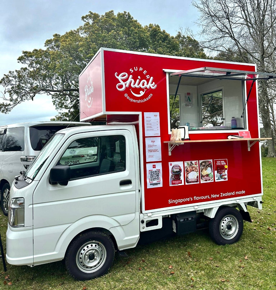 A white mini food truck with a bright red cargo box, parked on grass outdoors. The side of the truck features a large red panel with the words 'Super Shiok' in white script, a serving window, menu posters, and QR codes.